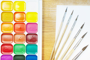 Watercolor paints, brushes and blank sheet of paper on wooden table. Set of artist tools for painting, concept for art and hobby. Top view