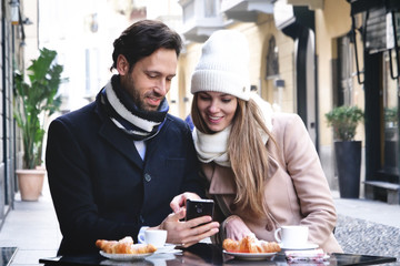 A loving couple is having breakfast in a bar while sitting outdoors and smiling as they look at the phone. Concept of: love, valentine's day, breakfast and happiness