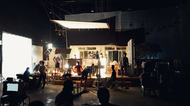 Silhouette Group Of People Working In Big Production Studio For Shooting Or Filming TV Commercial With Highly Quality Digital Video Camera And Professional Lighting Set And Prop.