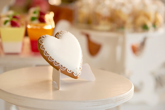 Close-up of a delicious heart-shaped glazed cookie standing on the wooden stand near the candybar with different desserts like yellow cupcakes and red jellies. festive choice gor wedding celebration.