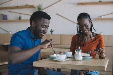 smiling african american couple at table with cups of coffee and desserts in cafe