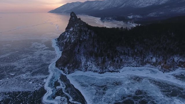 Sunset  deep cracks Lake Baikal. Unique Transparent blue smooth ice. High dark mountain rock snow. Cinematic Best Russia North Arctic. Winter season landscape orange colorful sunrise. Aerial Above