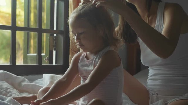 Young mother sits on the bed and combing her daughter in slow motion.