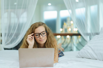 Happy young woman working on computer while laying at her working place in bed