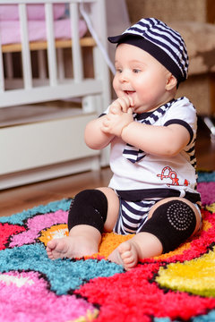 A pretty baby in a striped shirt and hats seated on the mat in the room