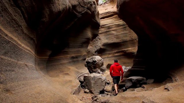 Young man on red hoodie walking through fascinating flowing shapes walls canyon in Gran Canaria, Spain. Hiker on adventure exploring eroded sandstone rock formation passageway. Unique nature landscape