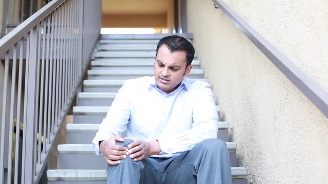 Closeup portrait, young handsome man in blue shirt, gray pants sitting on stairs in severe knee pain, isolated outside background. Negative human emotion