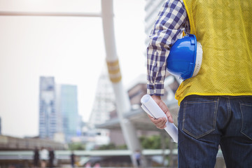 Engineering and construction concept. Closeup engineer with blueprint and helmet standing on site, blurred high building in city, with free copy space.