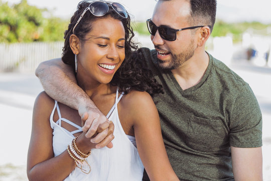 Content stylish African-American woman with handsome man in sunglasses embracing in happiness on background of coastline.