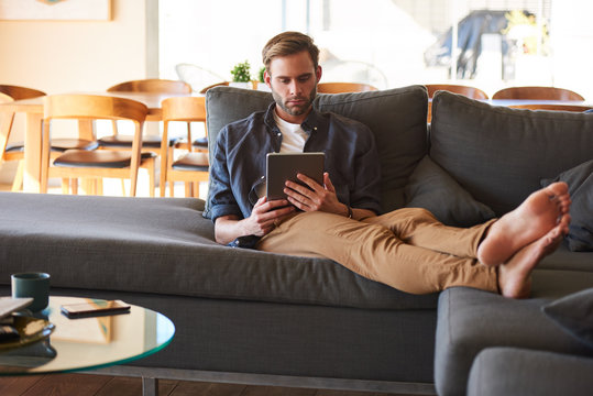 Attractive young white man sitting comfortably on his great couch with his feet up while surfing the internet on his electronic tablet to keep track of the latest news from around the world.