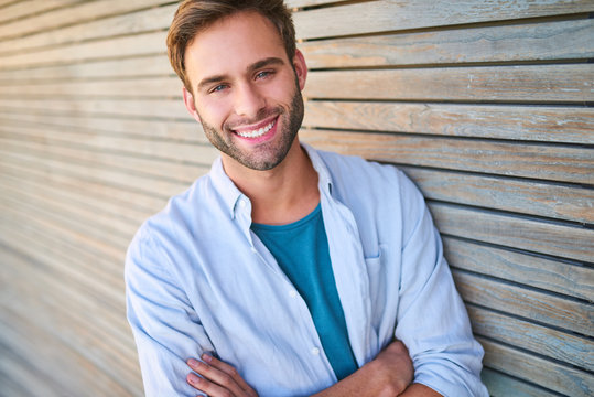 Tightly cropped image of handsome young male in his mid 20s smiling at camera with his teeth showing and his arms crossed while leaning against beautiful wooden cladding outdoors.
