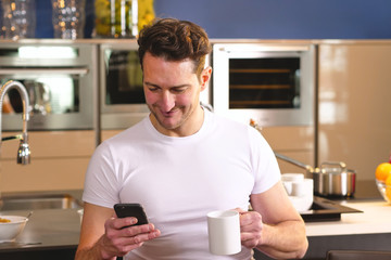A man in the kitchen while having breakfast and sipping tea or milk in the cup sends a message or calls with the tablet and smiles. Concept of: social network, message, technology.