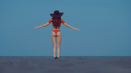 Back view of beautiful girl in bikini and hat relaxing in the sun during summer holiday on the sandy tropical beach over sea and sky background