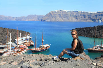 Fototapeta na wymiar Young woman enjoys view of excursion boats at small port on volcano of Santorini