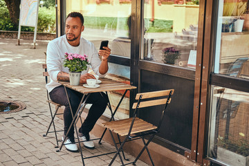 A fashionable African-American bearded male sitting near a coffee shop with a cup of coffee, using a smartphone.