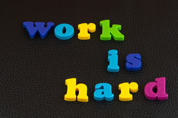 Work is hard  written in a mix of colourful plastic letters  on black leather. 
