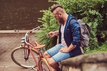 Fototapeta na wymiar Back view portrait of a bearded male with a haircut dressed in casual clothes with a backpack, sitting in a city park, using a smartphone.