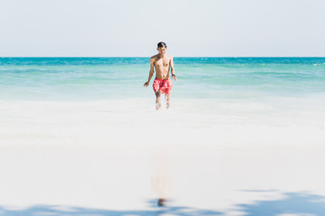 Fototapeta na wymiar Young man on beautiful sunny white sand beach on Ko Samet, Thailand: Asian man in red swimming trunks walks from turquoise sea waters to the wide empty shore, happy & wet after swim. Wide copy space