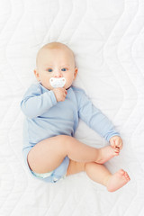 Cute baby boy with pacifier in blue onesie lying on bed