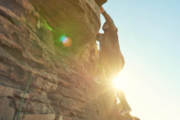 African guy climbing a near vertical rock face with clear blue skies and flare from the sun shining...