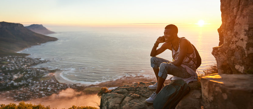 Panoramic image of a young black man sitting on the top of a mountain talking on a mobile phone during an amazing sunset, with a beautiful view of the ocean and cityscape in the background.
