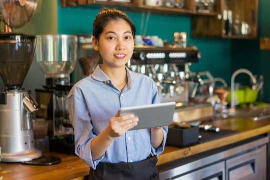 Cheerful Asian coffee shop employee using modern device and looking at camera. Positive confident small business owner working in coffee house. Entrepreneur concept