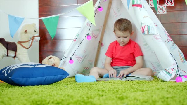 Little boy is reading book in his self made wigwam in playroom