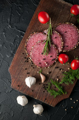 Smoked salami on a black table and a brown board. Sausages with rosemary, garlic and pepper and tomato cherry, dill, parsley.