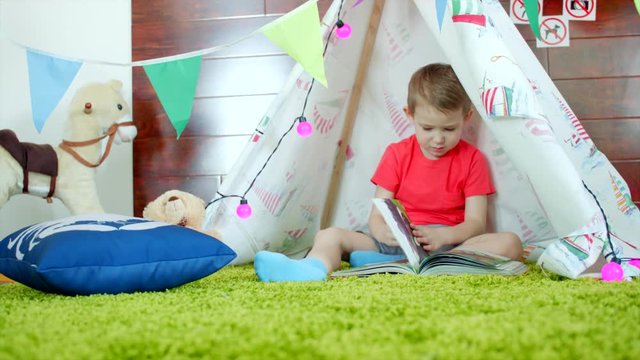 Little boy is reading book in his self made wigwam in playroom