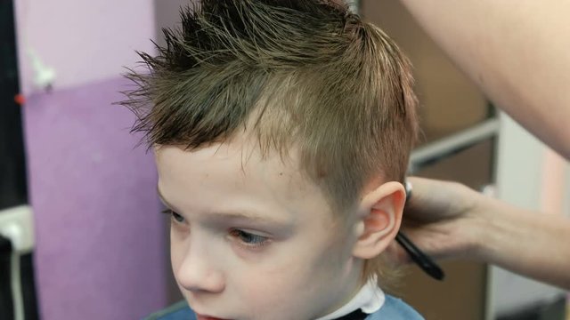 Barber's hands combs and cutting blond short boy's hair with scissors. Closeup boy's face.