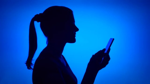 Silhouette of young woman using mobile on blue background. Female's face in profile browsing, reading, chatting online with friends on cellphone. Black contur shadow of teenager's half-face