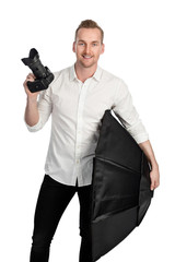 Smiling photographer standing against a white background holding a digital camera and a softbox.