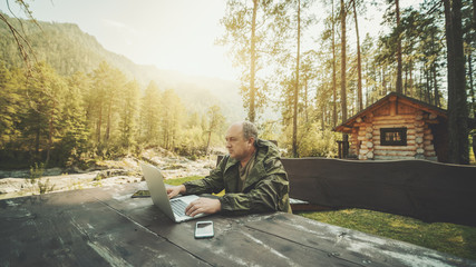 Adult partly bald hunter is sitting outdoors near his shack at the wooden table and working on his...