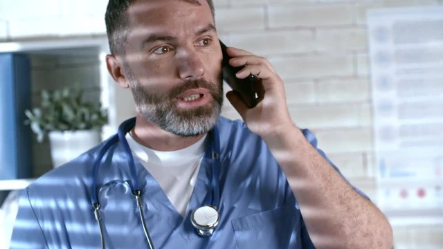 Handheld medium shot of bearded male doctor wearing scrubs and stethoscope around his neck talking on mobile phone in private practice office
