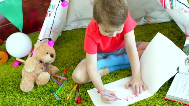 Little boy is writing MAMA in the drawing album in the playroom