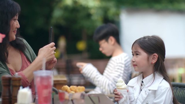 Cute Asian little girl sitting with ice cream at cafe table outdoors and posing while mother photographing her with smartphone