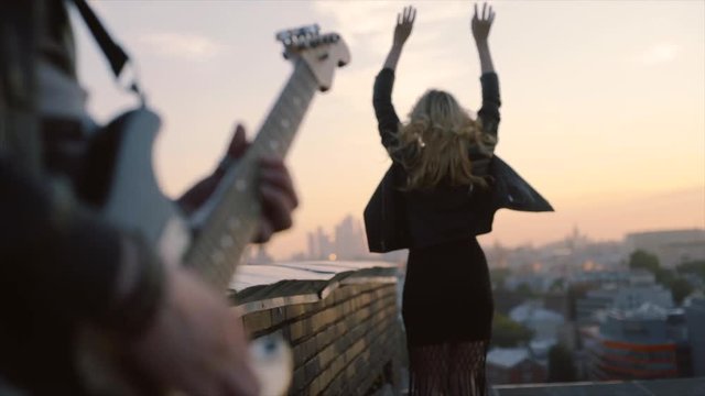  young women in rock n roll style clothes walking on the roof enjoying wind, man playing on guitar close up hands and instrument at blurred foreground 