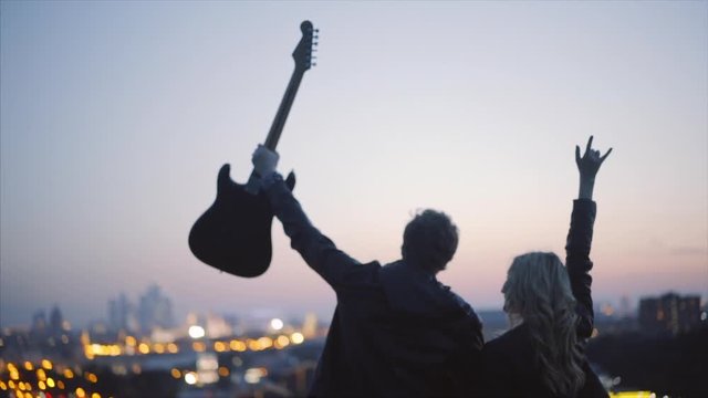 couple in rock-n-roll style standing on the roof with guitar in hand expression makes the rock n roll gesture in evening time at blurred city lights background slow motion shot .