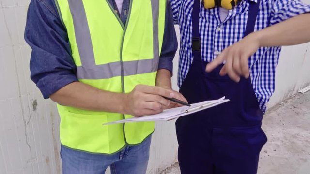 Medium shot of two construction workers in hard hats, one holding clipboard with documents, discussing project details and pointing at something