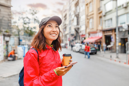 Cheerful young woman wearing pink jacket smiling and holding her smartphone in the busy city street with cars and drinking take away coffee in paper cup.