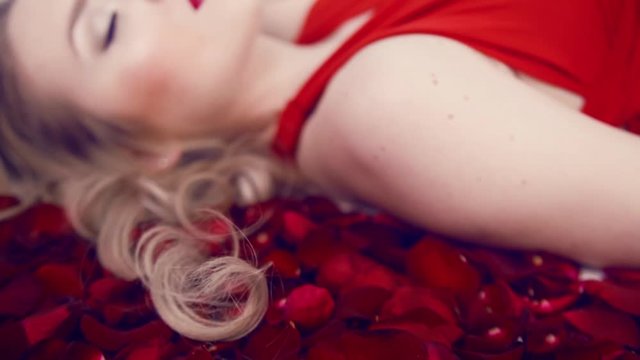 Beautiful blonde girl lies in a lot of red roses. Red lips and gentle makias. Girl in a red dress. Valentine's Day. Light studio. Lies with her eyes closed, touches her hair.