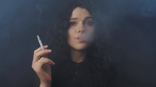 Portrait of young brunette woman with curly hair is smoking a cigarette and looking at the camera in dark smoked room