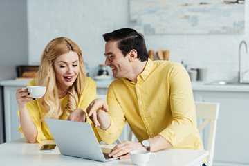 Happy couple drinking coffee and talking by table with laptop