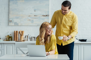 Attentive boyfriend giving coffee to woman working by laptop