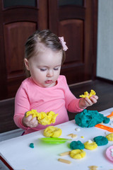 A 1.5 year old girl sits at a table and plays with a colored dough, on the table lie tools, molds and pasta for decor