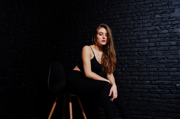 Obraz na płótnie Canvas Handsome brunette girl wear on black, sitting and posing on chair at studio against dark brick wall. Studio model portrait.