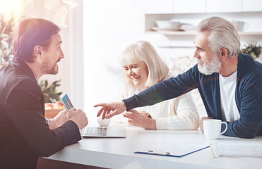 Choose the options. Cheerful professional insurance agent showign the information on laptop while meeting with elderly loving couple in the kitchen