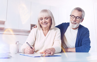 Fototapeta premium Place for signature. Cheerful delighted old couple sitting in the kitchen and signing the insurance contract while expressing positivity
