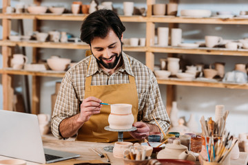 smiling potter painting ceramic jug in workshop with laptop