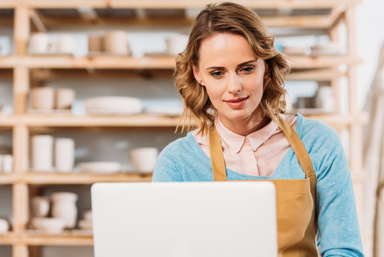 beautiful woman in apron using laptop in pottery workshop